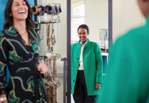 A woman trying on a green coat and smiling at her reflection in the mirror, while another woman, dressed in a patterned dress, laughs.