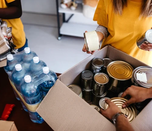 People packing canned goods and bottled water into cardboard box.